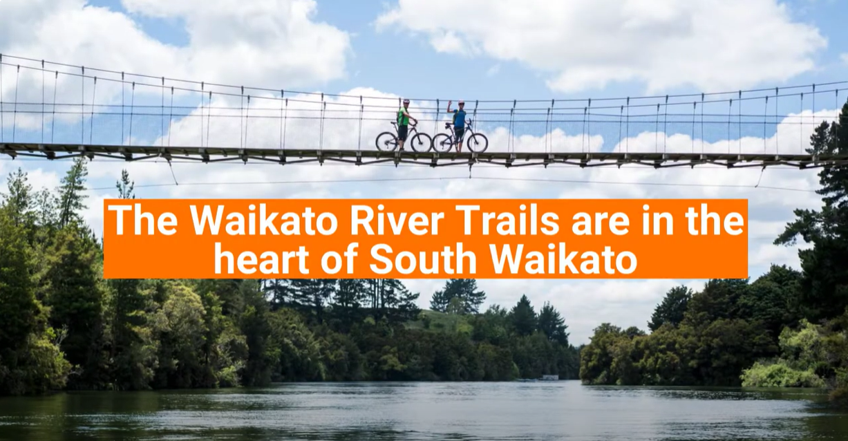 The Waikato River Trails are in the heart of South Waikato featuring two mounted bikers waving from a swing bridge over the river.