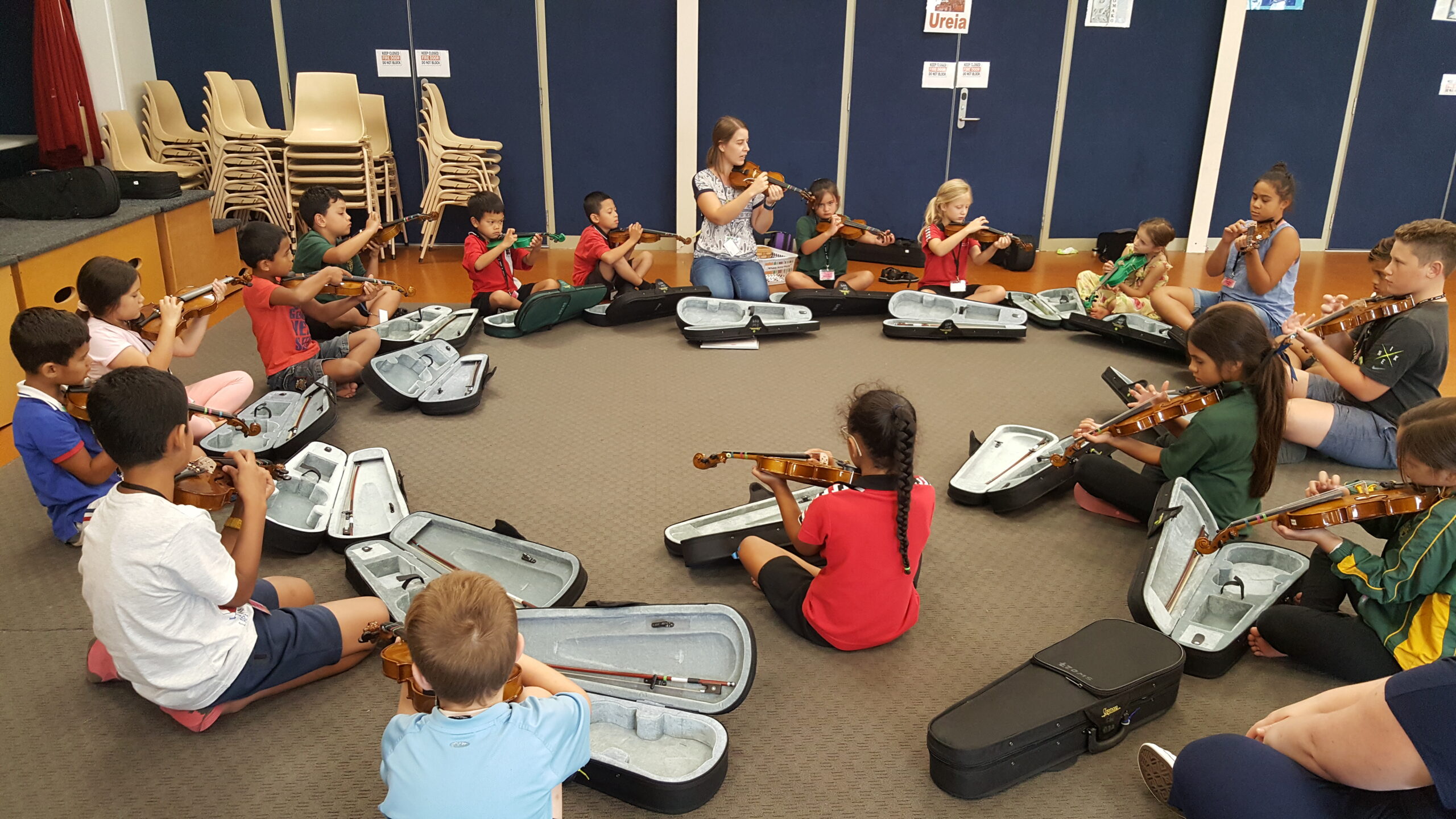A group of children sitting in a circle on the floor holding violins, with a teacher explaining in the front middle.