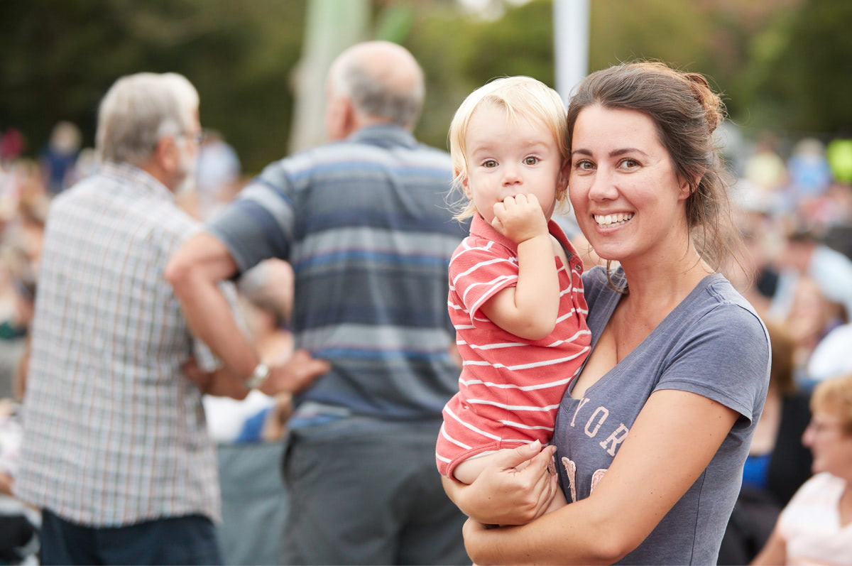 Mum holding child with a blurred crowd in the background at a Hamilton Gardens Arts Festival event
