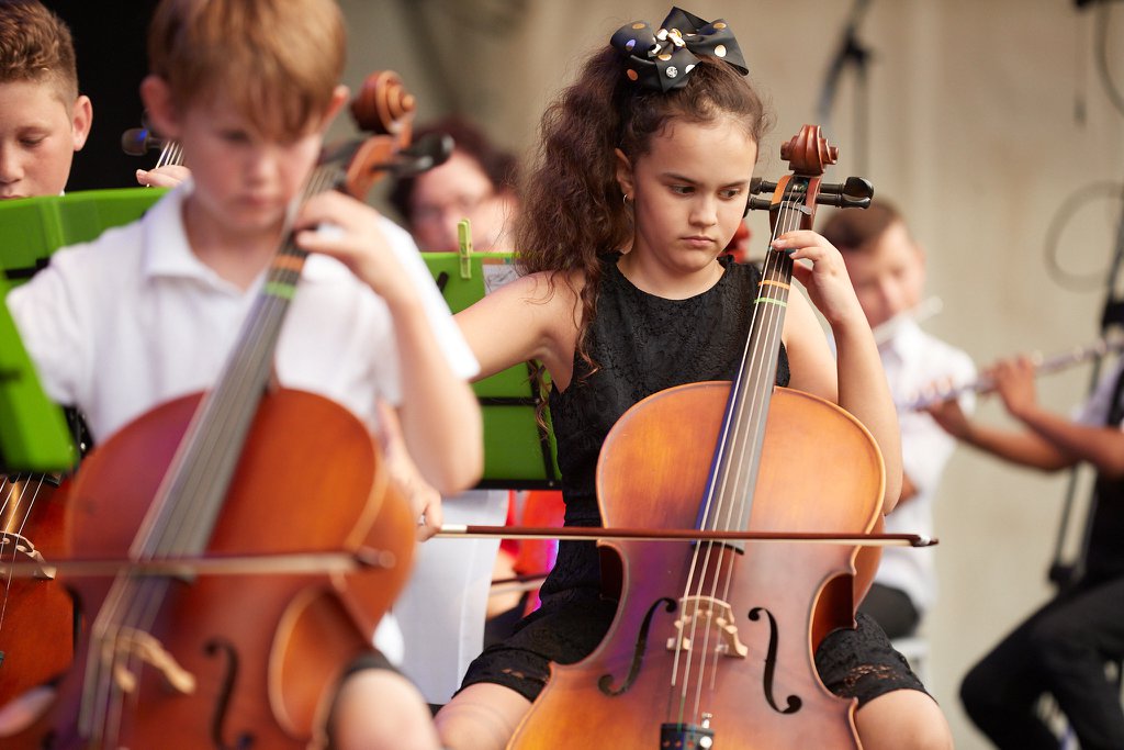 School kids doing an orchestra performance, with cello players in the forefront and some flutists in the background