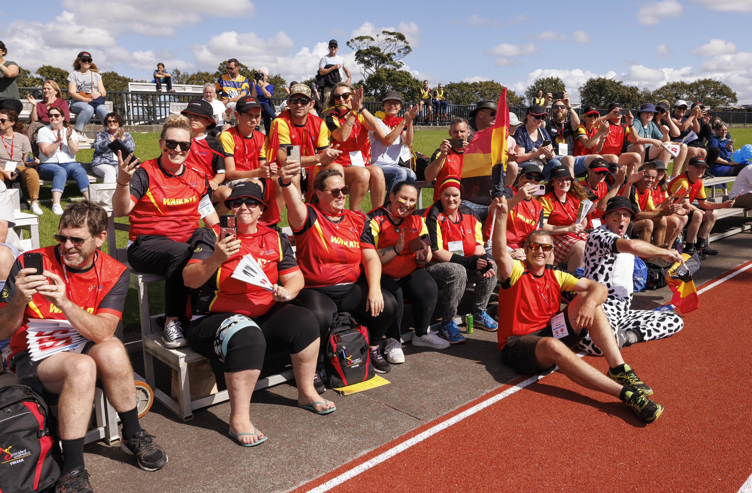 Crowd of Waikato supporters cheering and smiling on the sideline benches of a running track
