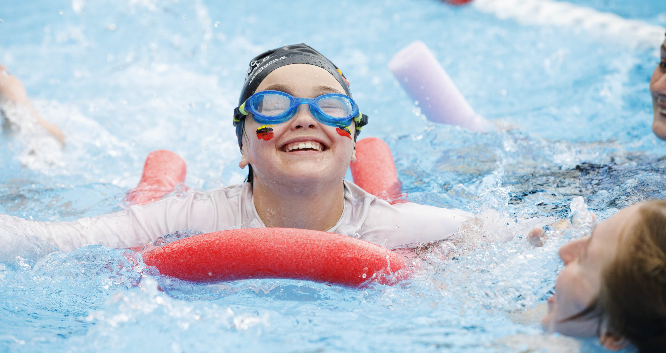 Girl with Waikato colours face painted on her cheeks swimming with a pool noodle wearing goggles and a swim cap