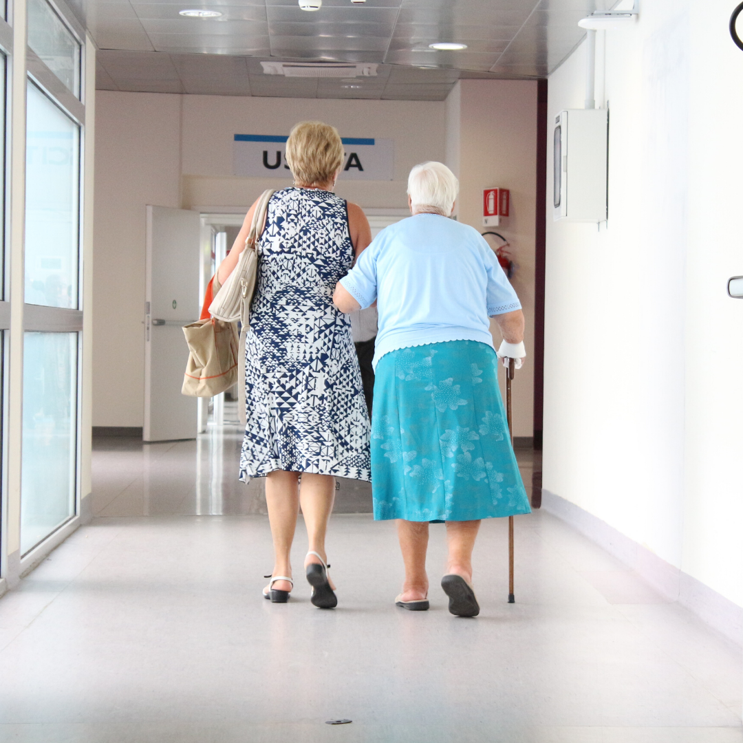 Elderly lady walking down hospital hallway using a walker getting help from another woman.