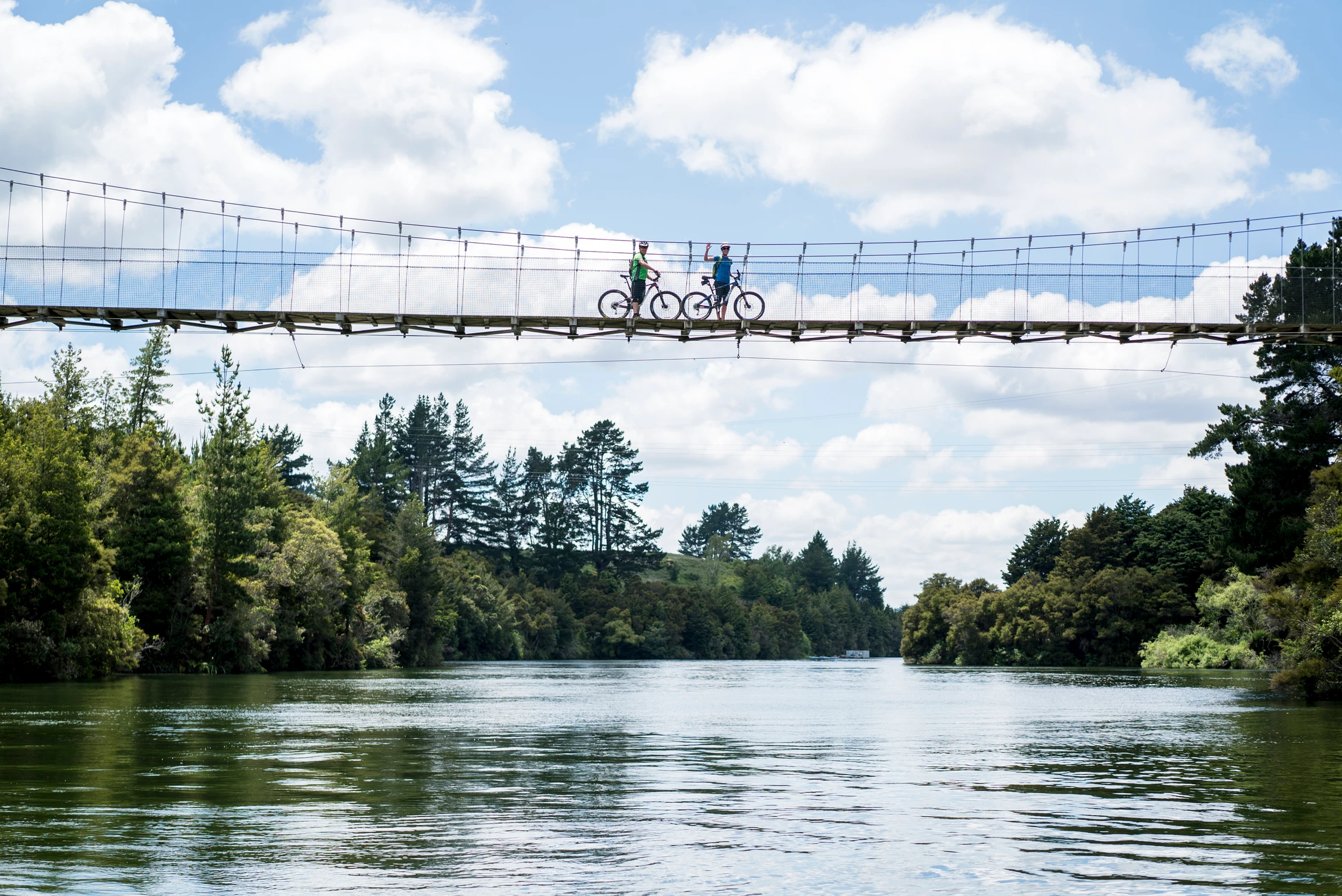 Two mounted bikers waving from a swing bridge over the river.