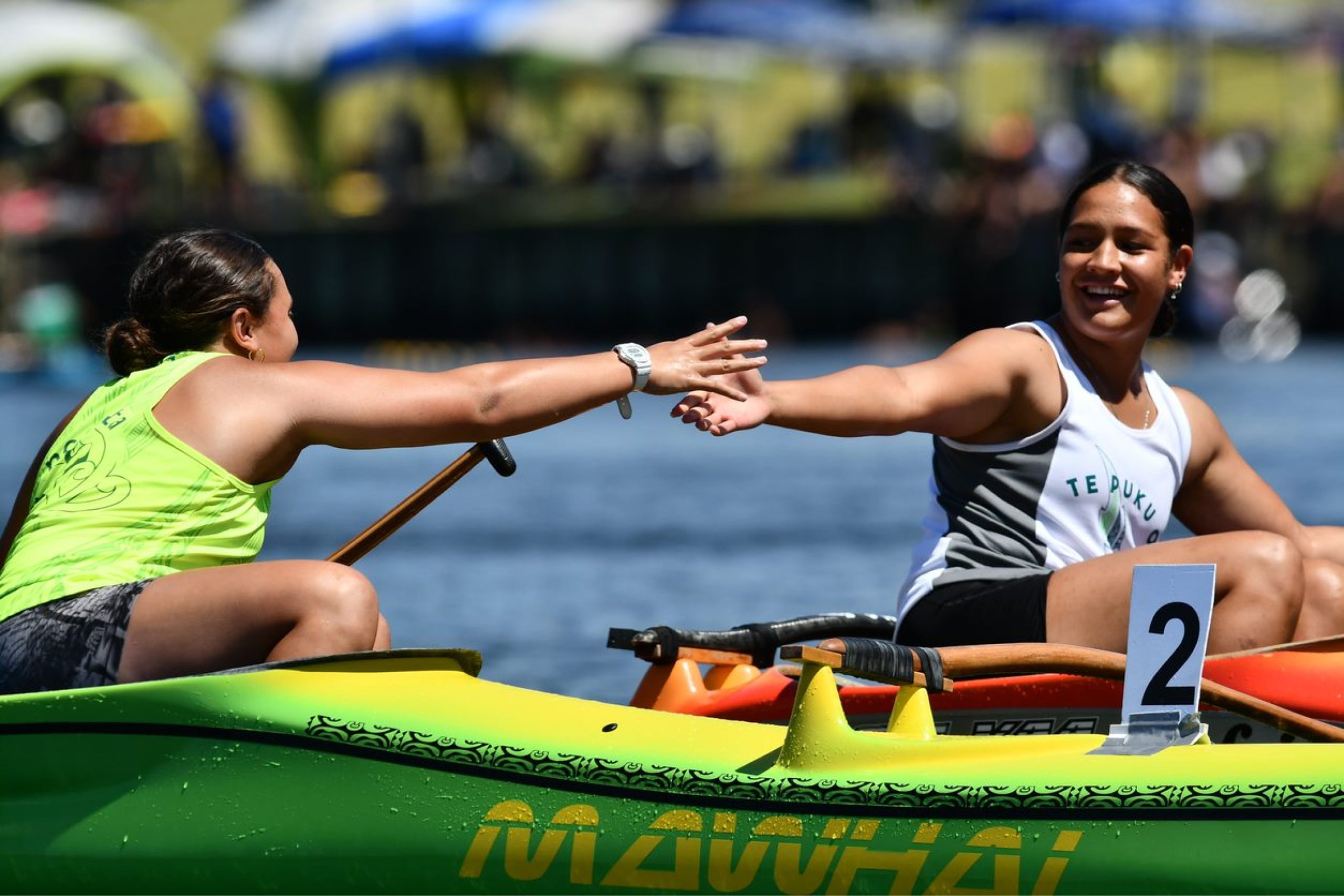 Two young woman reaching out to each other from their waka at a waka ama event